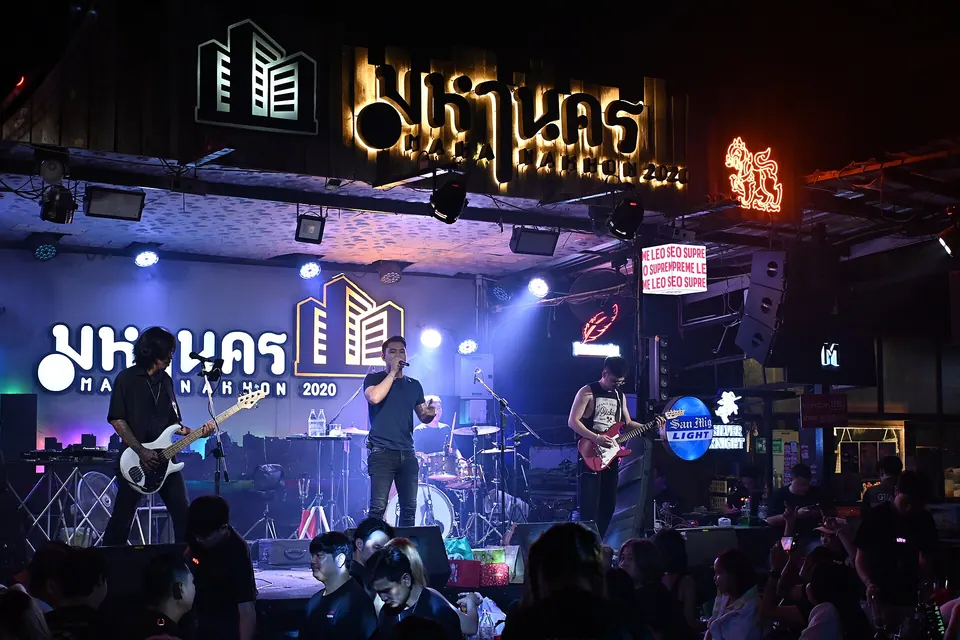Photo: a four-piece band on the lit stage at Mahanakhon under its Thai name sign, the crowd at tables in front